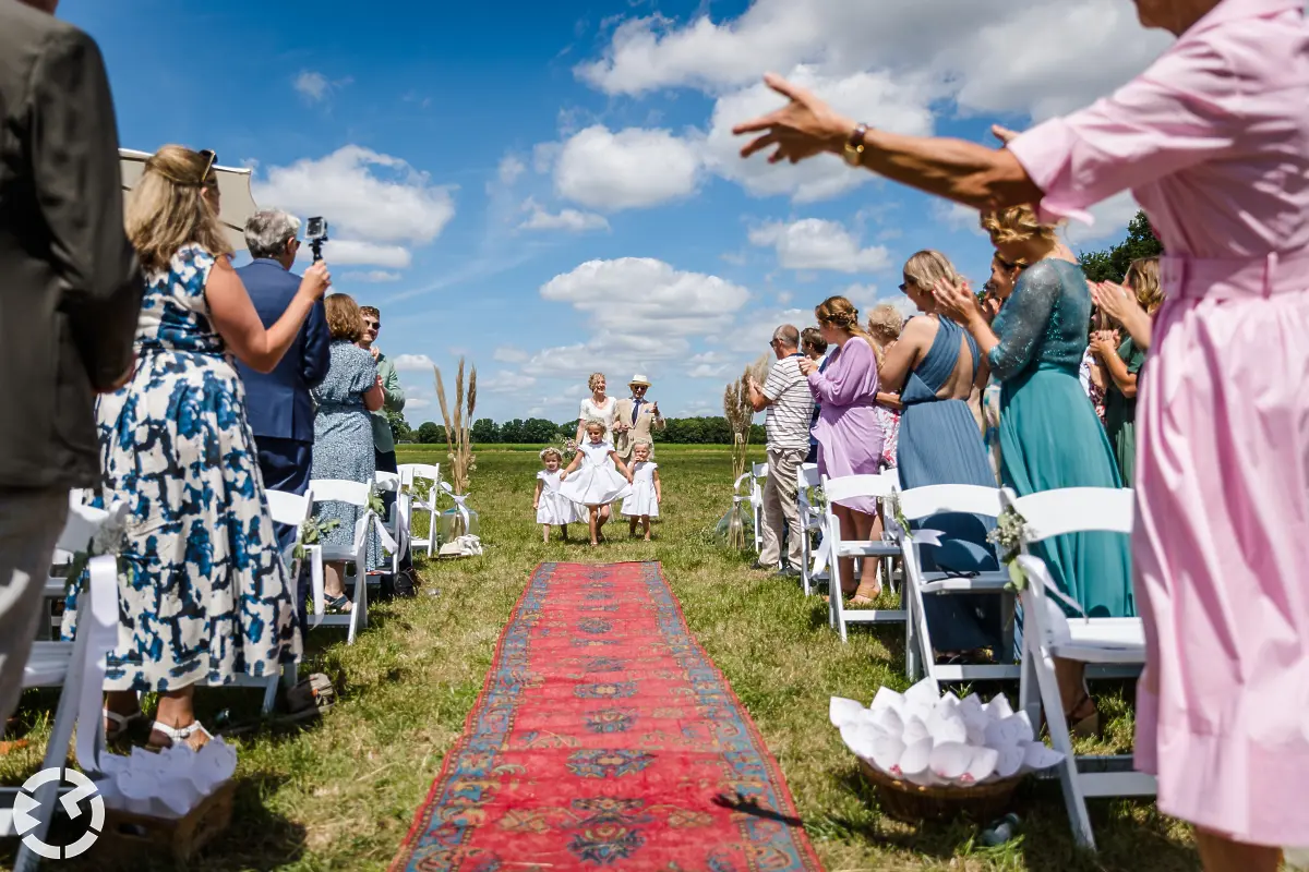 Bruid komt ceremonie op met bruidskinderen en vader over een lang perzisch tapijt in een weiland.