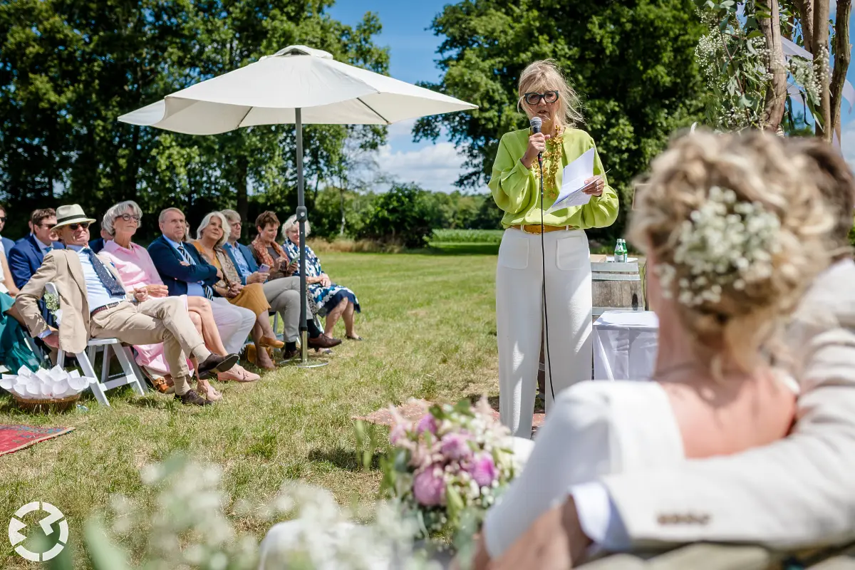 Bruidspaar wordt door de trouwambtenaar toegesproken en gasten onder een parasol luisteren aandachtig.