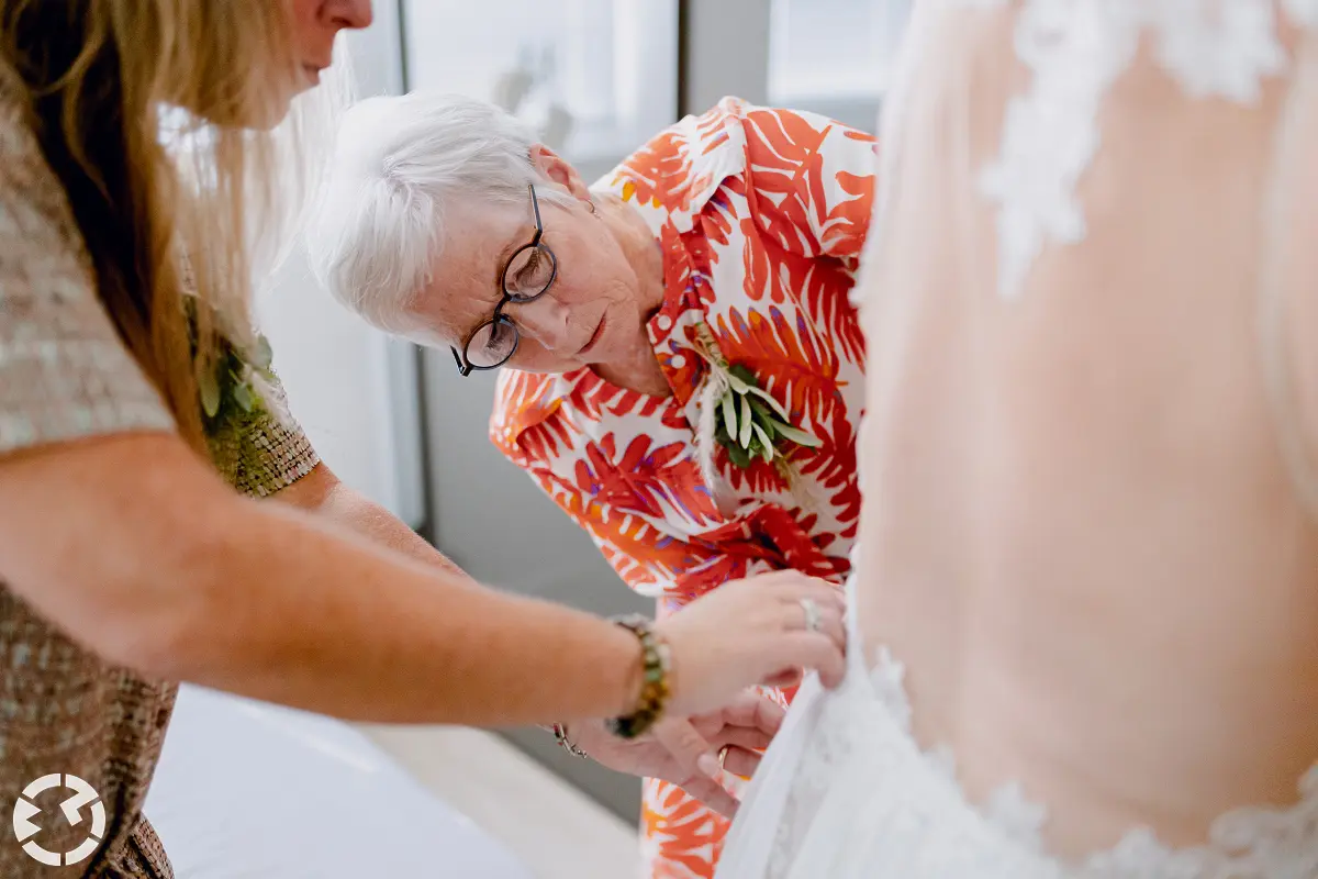 Vrouw met wit haar kijkt aandachtig naar de zijkant van de trouwjurk die de bruid net aan heeft getrokken.