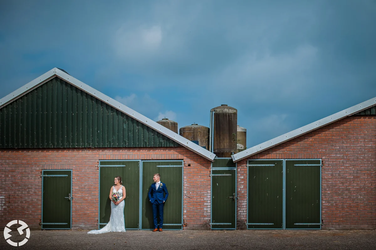 overzichtsfoto van een boerderijenstal waarbij het bruidspaar beide voor één deur staan.