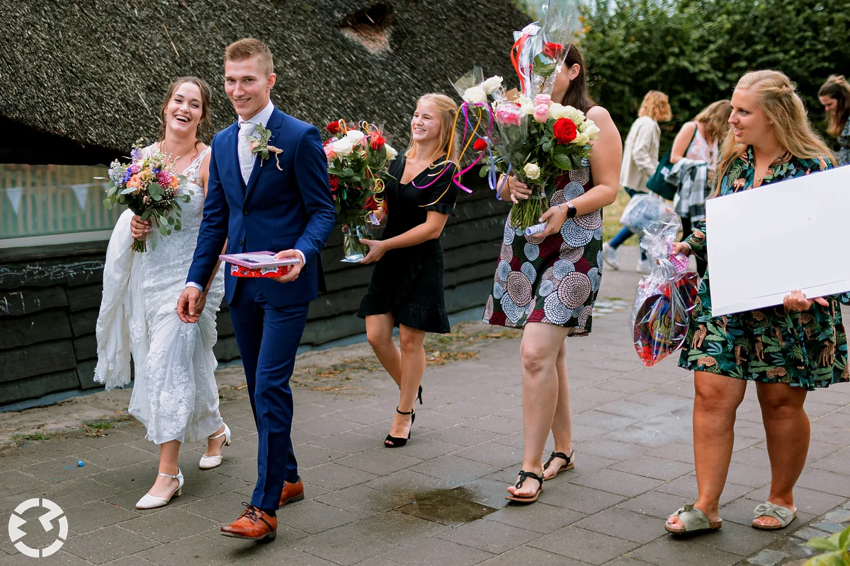 bruidspaar loopt weg bij een boerderij met bloemen in hun hand gevolgd door vrouwen die ook bloemen dragen