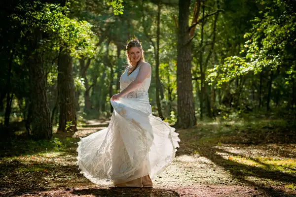 Bruid zwaait met haar jurk tijdens de fotoshoot in het zonlicht van een bos.